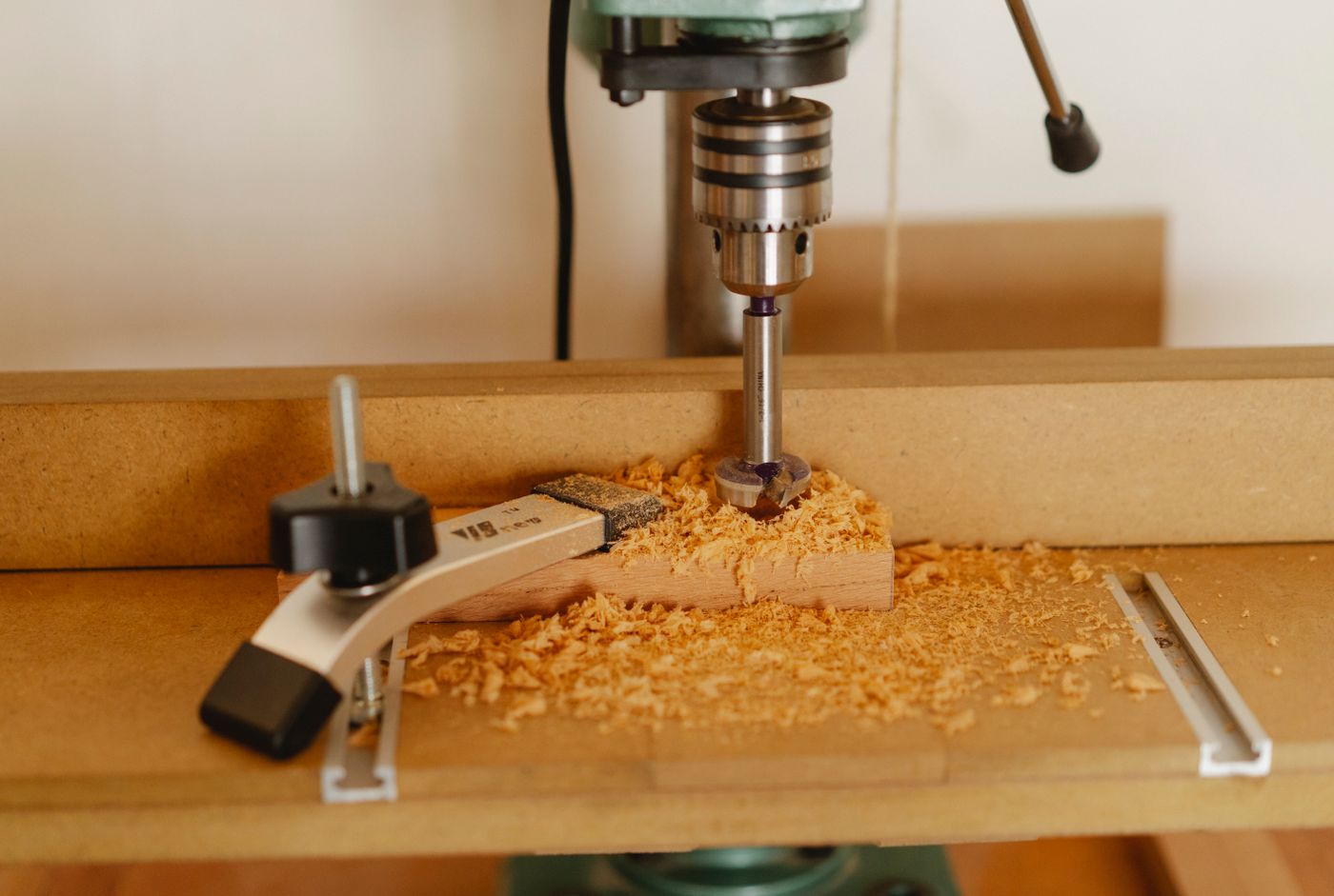 A drill press above timber held by a clamp on a workshop table
