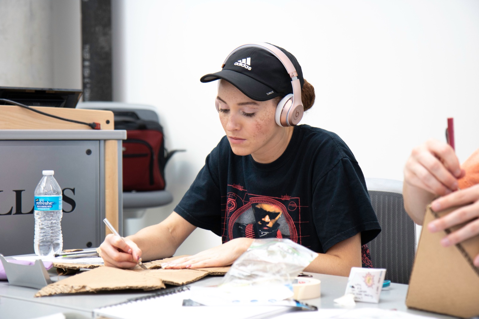 A person working on cardboard at a clear classroom table with a cutting tool and project materials