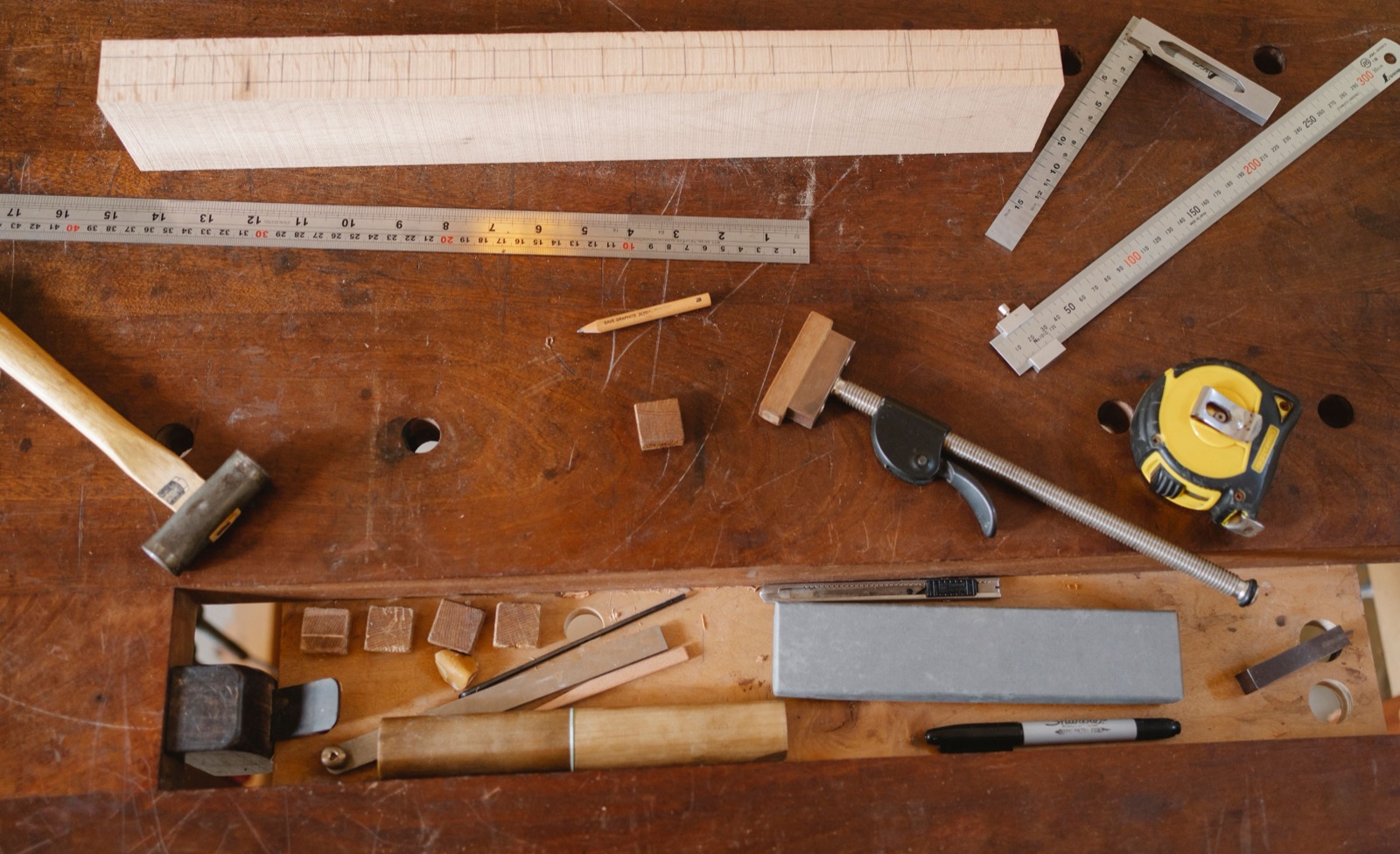 A woodworking bench with a board, rulers, square, clamp, pencil, and other tools laid out for a project